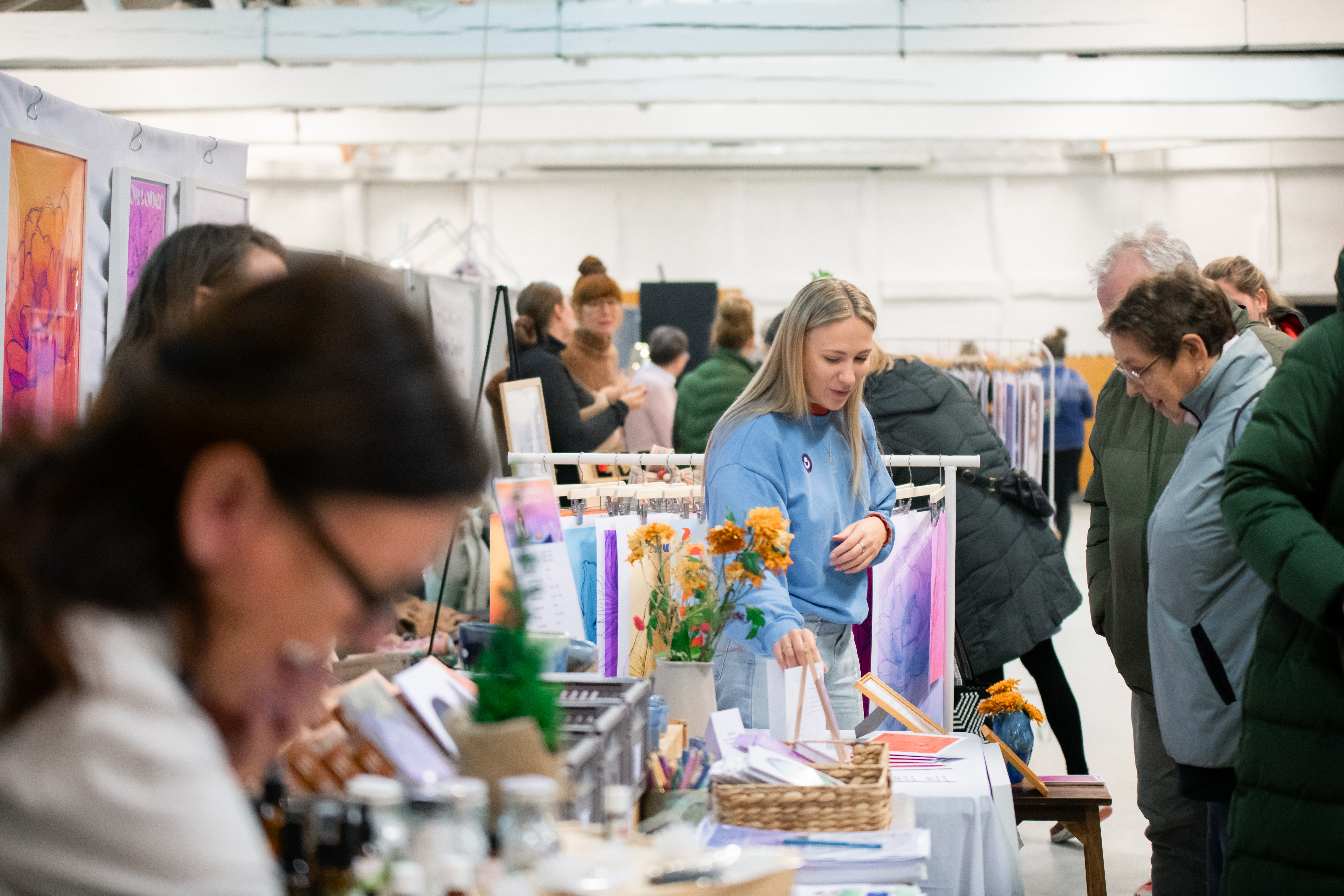 Person sløret i forgrunden, i mellemgrunden står en kvinde og viser noget frem til nogle markedsgæster. Markedet fortsætter i baggrunden. 