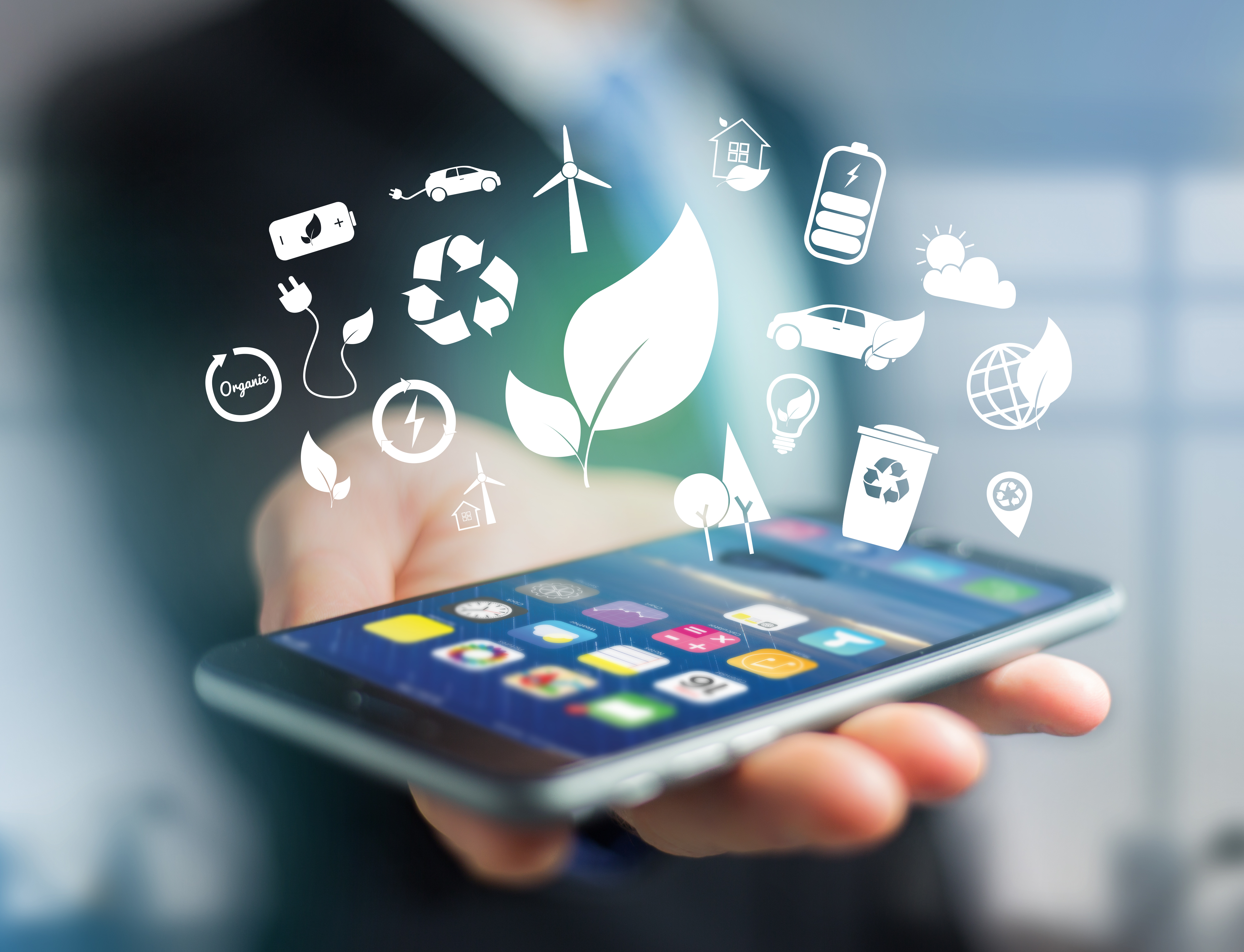 Nærbillede af en mobiltelefon, der ligger i hånden på en person klædt i mørkt jakkesæt og slips. Over telefonen sværer hvide ikoner som et blad, en vindmølle, en skraldebøtte med genbrugstegnet.