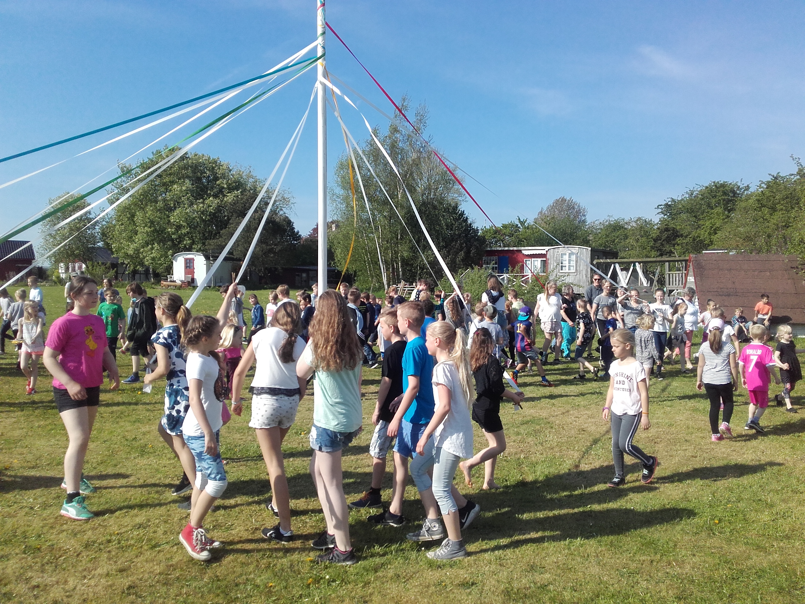 Børn og unge danser rundt om pæl med farvede bånd. 