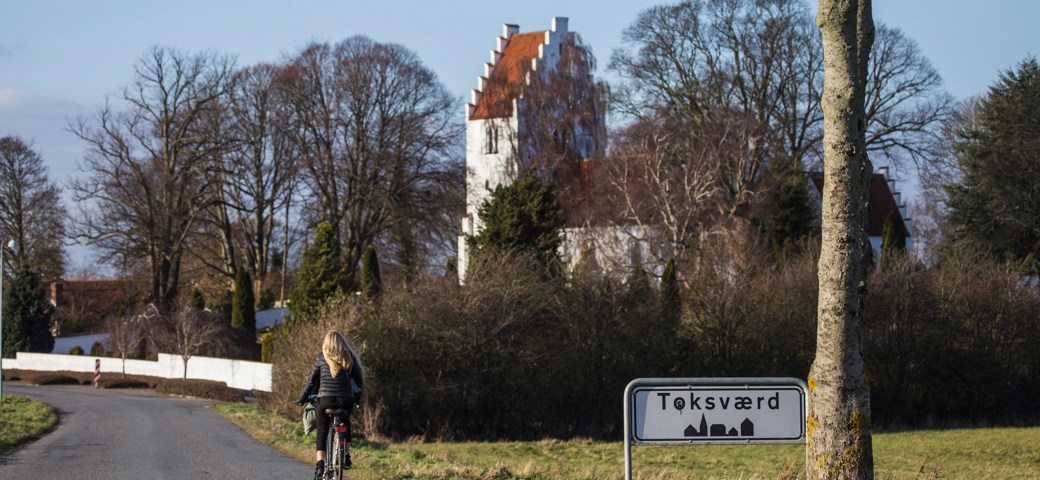 En pige er cyklet forbi byskiltet på vej ind til Toksværd med kirken på hendes højre side