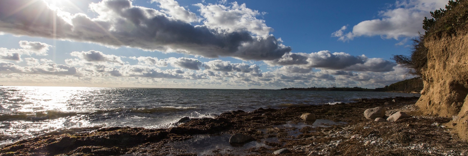 Solen skinner igennem et let skydække imens bølgerne slår ind på kysten ved Karrebæksminde