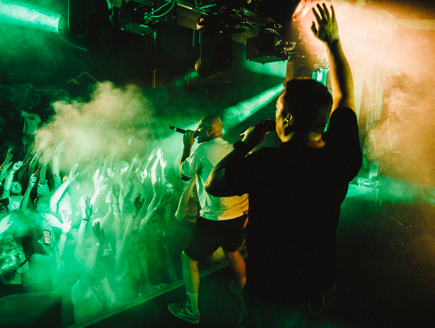 Koncert billede taget fra scenen. Kunstnere i forgrunden, grønt skær og røg i luften. Publikum har hænderne i luften. 