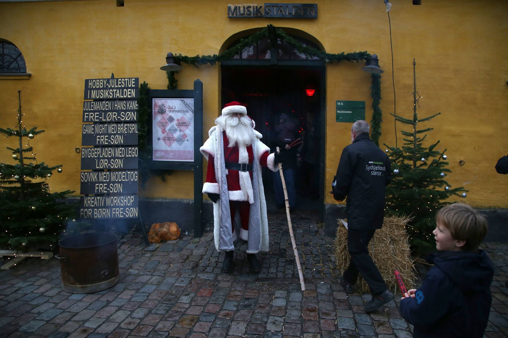 Julemanden holder en gren i hånden foran en gul bygning, Musikstalden på Grønnegades Kaserne Kulturcenter. 