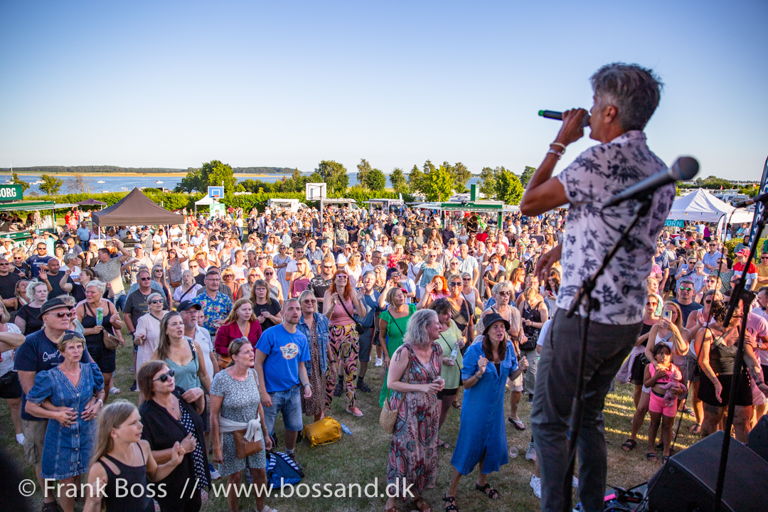 Mand står med mikrofon og synger. Publikum står foran scenen og synger med. I baggrunden ses Karrebæk Fjord. 